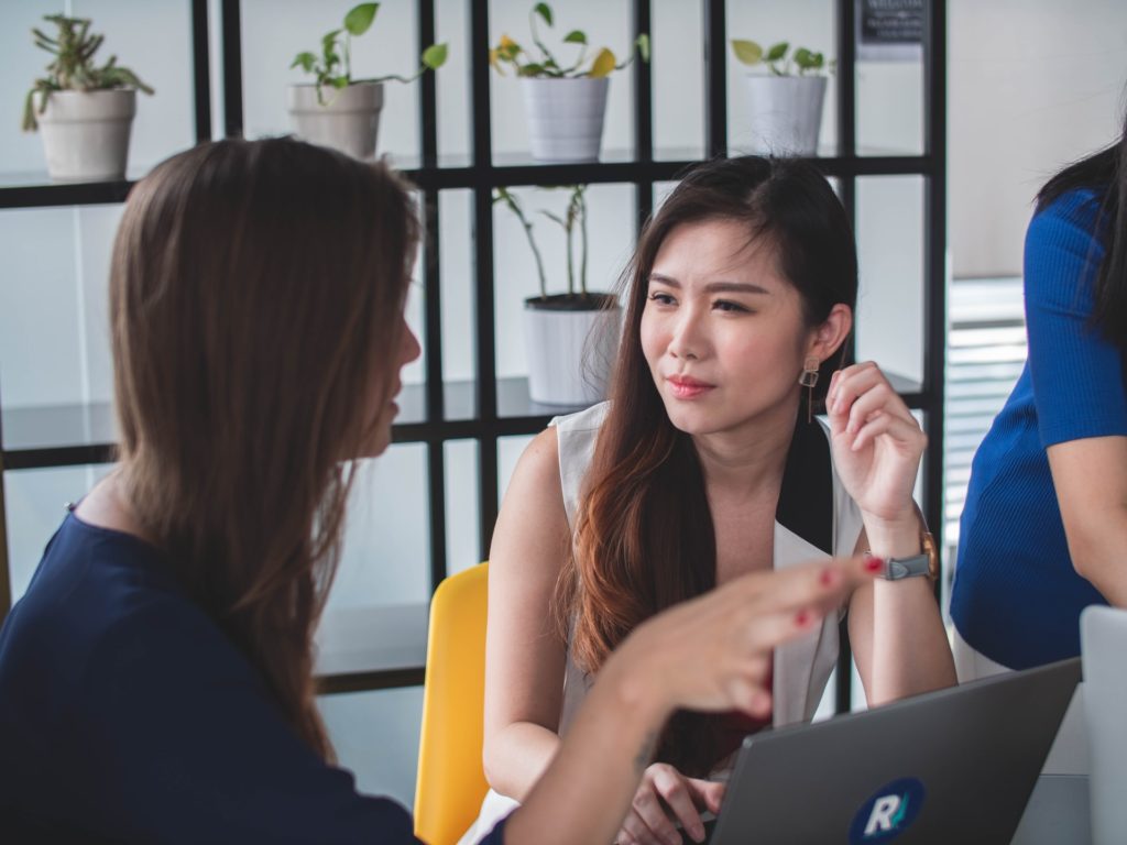 Two women in a conversation, one is talking and gesticulating, other listens keenly with her hand above a laptop