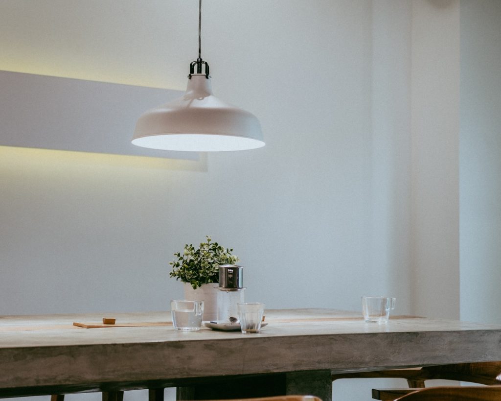 Stout, wooden table with a small potted plant and couple of cups on it. Lamp hangs above it, providing bright, but warm light